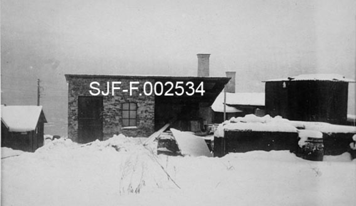 Fra de nye produksjonsanleggene Schwencke & Co's Eftf. bygde på Børsholmen i Asker i 1924.  Schwencke & Co. var et firma som produserte og forhandlet bek og andre tjærebaserte produkter.  Nettopp i 1924 ble mye av produksjonen flyttet fra Kongshavn sør for Oslo til Børsholmen, hvor den ildsfarlige virksomheten kunne drives i betryggende avstand i forhold til annen bebyggelse.  Dette fotografiet viser noen av bygningene på anlegget.  Midt i bildet ligger et hus med pulttak.  Under den venstre halvdelen av dette taket var det et rom med teglsteinsmurte vegger.  På den veggen som vendte mot fotografen var det ei dør og et støpejernsvindu.  Under den høyre delen av det samme pulttaket later det bare til å ha vært et åpent skur da dette fotografiet ble tatt.  Til høyre i bildet ser vi et lavt murverk (kanskje en uferdig bygning) med en diger, vertikalstilt stålsylinder inni.  Bakenfor skimtes et par andre tak og et par skorsteinspiper.  Fotografiet er tatt på snødekt mark. 