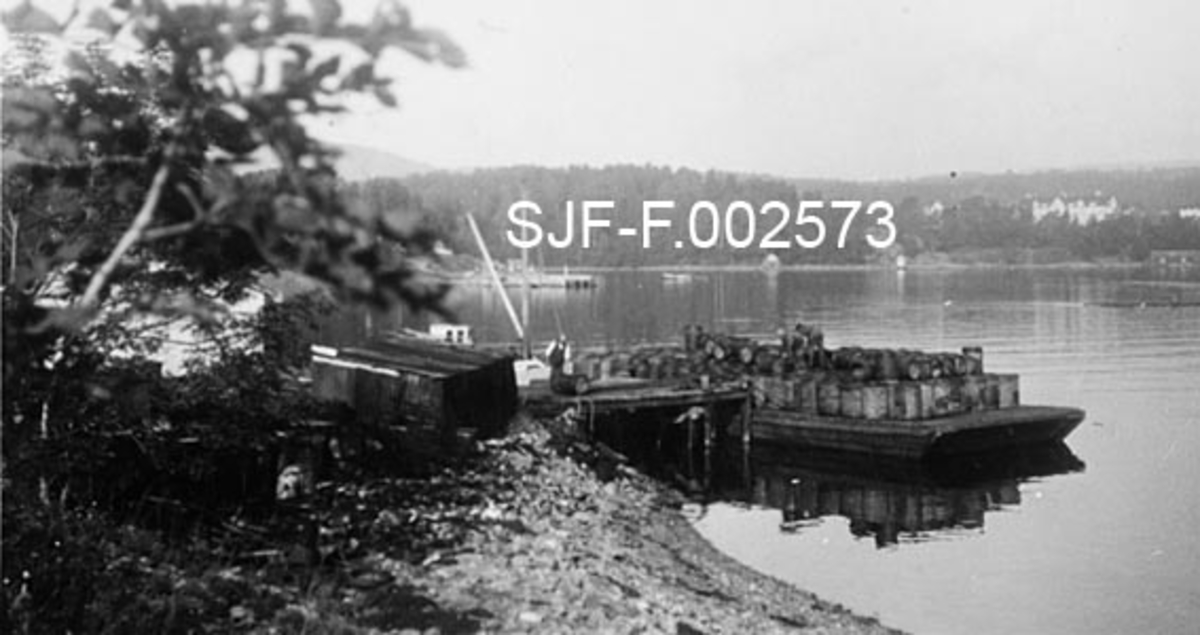 Fra kaia til Schwencke & Co's Eftf. på Børsholmen i Asker.  Fotografiet er tatt fra holmen, langs strandkanten mot kaia, hvor det lå en lekter (lastepram) med en mengde tønner, som enten skulle lastes eller losses.  Fire menn arbeidet på lekteren, en på kaia.  Bak sistnevnte skimter vi en båt som lå på baksida av kaia.  Inne på land lå et skur med pulttak.  Fotografiet ble tatt i 1941.  Schwencke & Co. produserte og forhandlet bek og andre tjærebaserte produkter. 