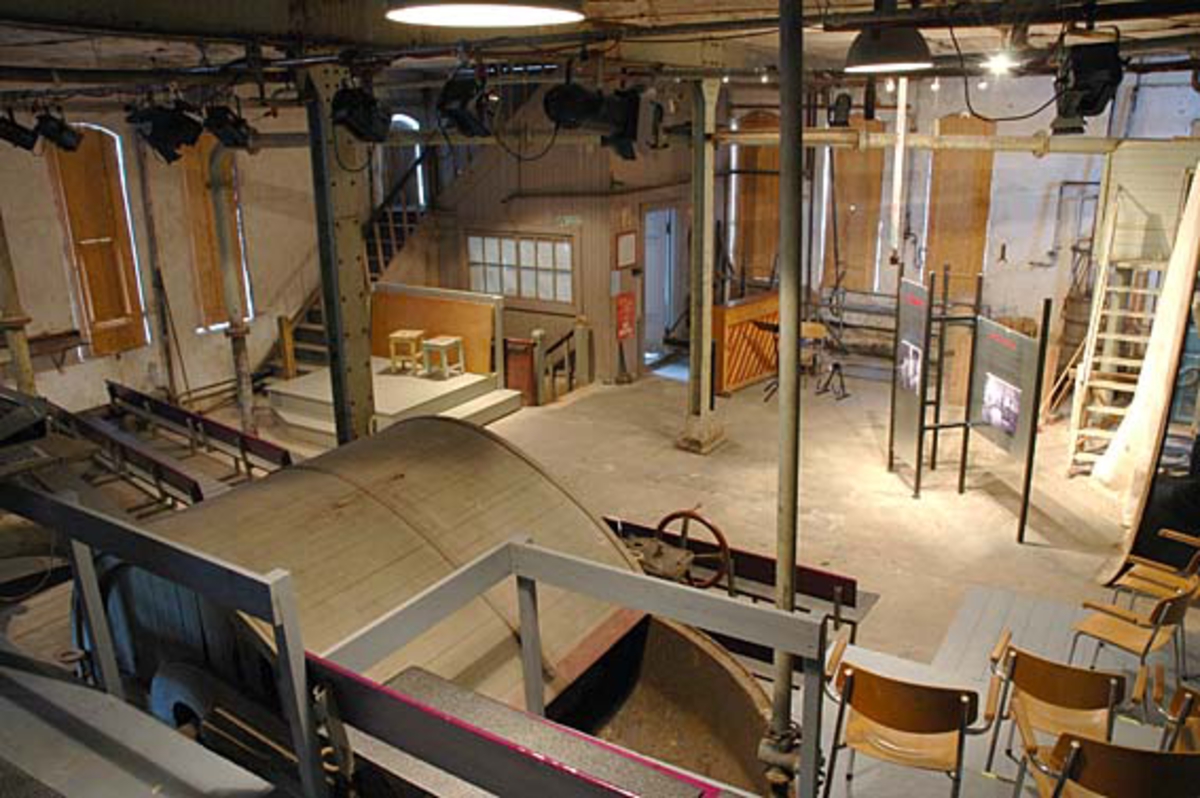 Hollenderiet på Klevfos Industrimuseum. Produksjonslokalet er blitt "Teatersal" for å dokumentere arbeidslivet før, med sang og teater. I taket er det oppmonter teaterbelysning, blandt autentisk belysning.  Løten. Ådalsbruk.
