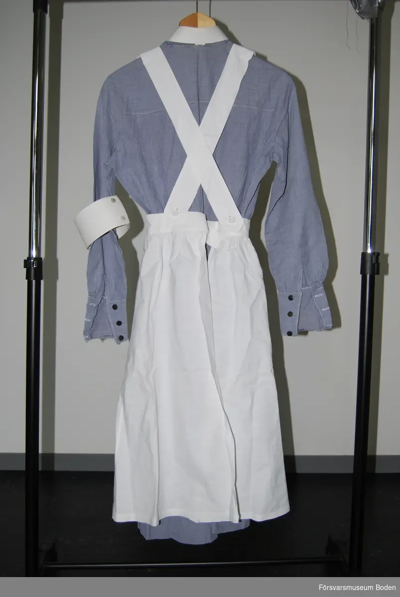 Smårutigt bomullstyg i blått och vitt. Klänningen användes tillsammans med vitt förkläde, mörkblått skärp, vit krage, brosch, armbindel samt på huvudet vit hätta, vilka utgjorde arbetskläderna för en sjuksköterska vid Röda Korset.