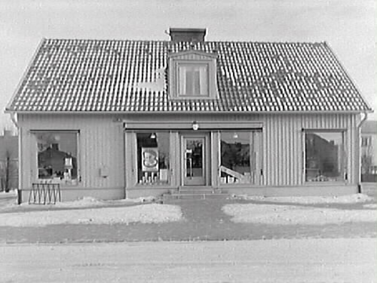 Kooperativa AB. Butiken låg i en fristående träbyggnad utmed Södra vägen, kv Gösen. Entrén ligger centrerad tillsammans med skyltfönster i ett svagt utskjutande fasadparti. Bilden är tagen på vintern, det ligger snö på marken.