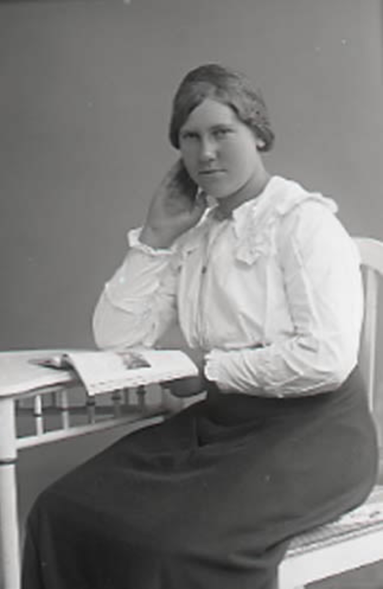 Ateljéfoto, sittande ung kvinna med skrift vid bord. Beställare: Evelina Karlsson, Vadkärr, Veddige.