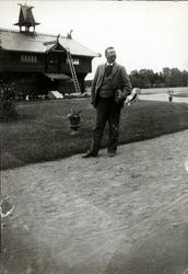 Portrett, ukjent mann, stående utendørs på tun foran trebygn