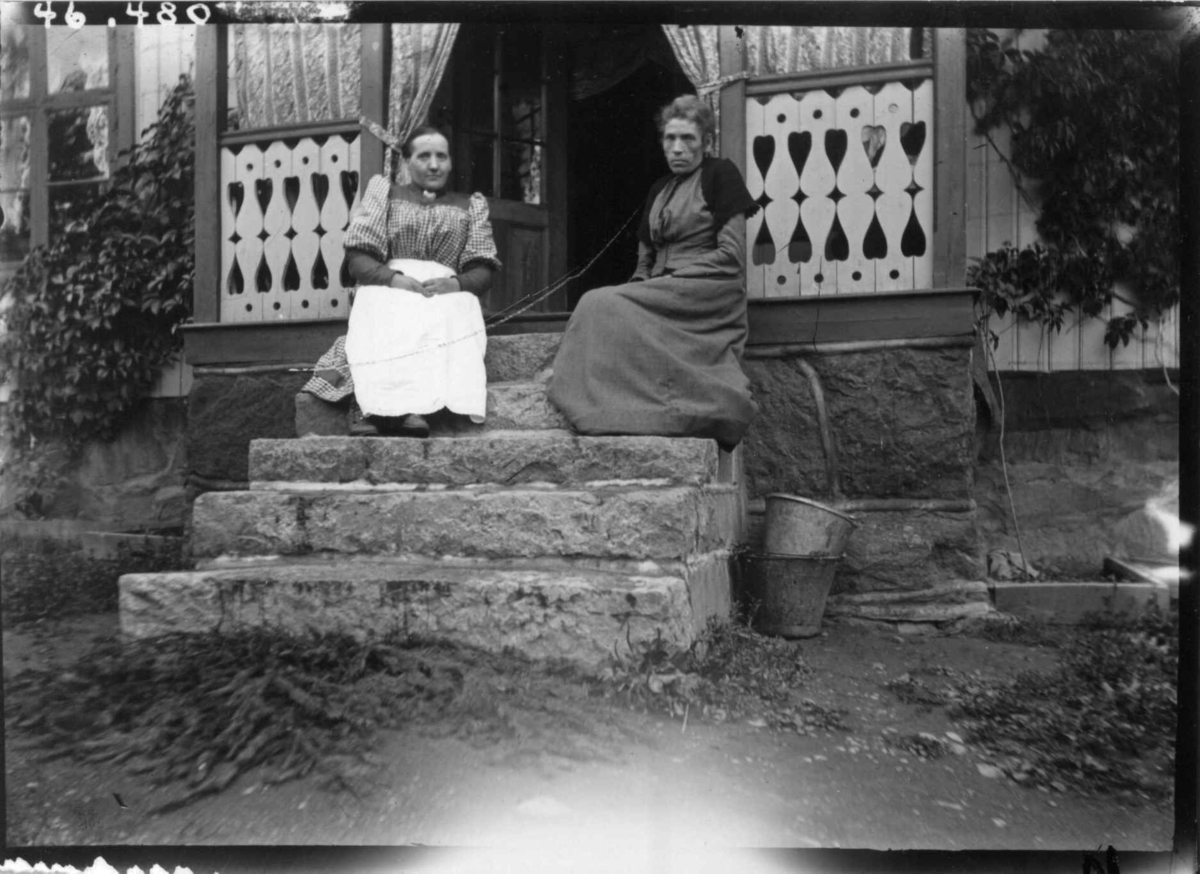 Drakt 1908-10. To kvinner sittende på en trapp. I lange kjoler. I bakgrunnen en veranda. 