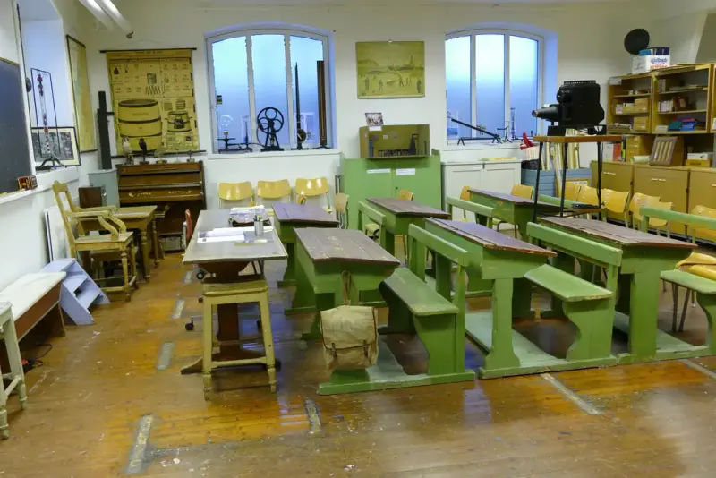 Bilde av skolepulter i treverk med gammeldags skolesekk og plansjer på veggene