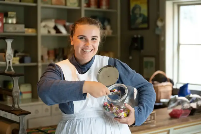 Smilende kvinne i iført blå kjole og hvitt forkle plukker drops fra et glass foran disken i Kolonialen i Norsk Folkemuseum.