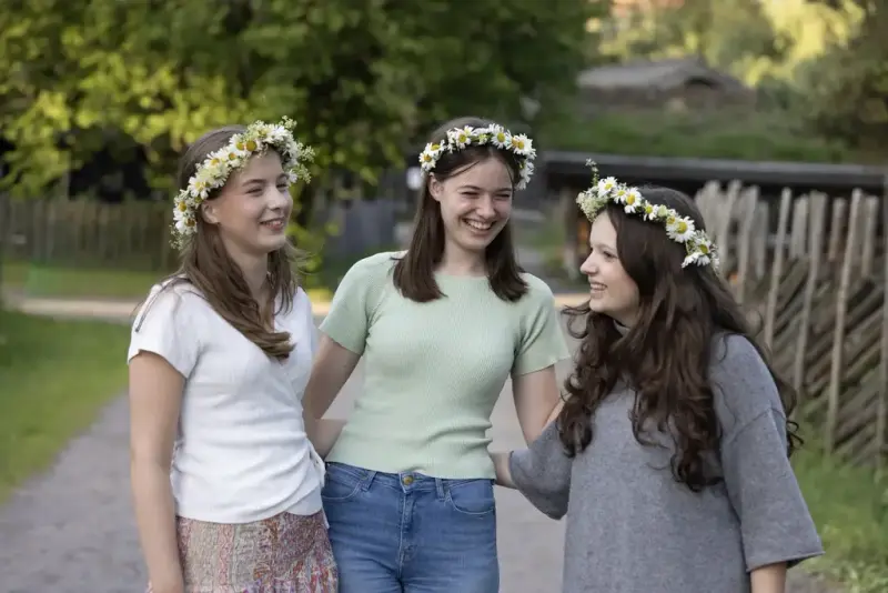 Tre unge kvinner med blomsterkranser smiler og koser seg på Sankthansfestivalen på Norsk Folkemuseum.